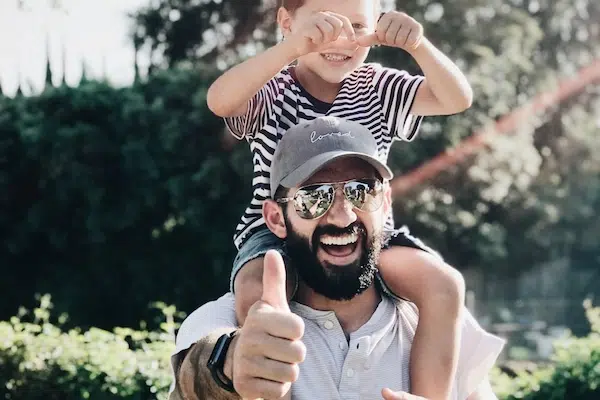 Smiling man wearing sunglasses and a cap gives a thumbs-up while carrying a child on his shoulders in a sunny outdoor setting.