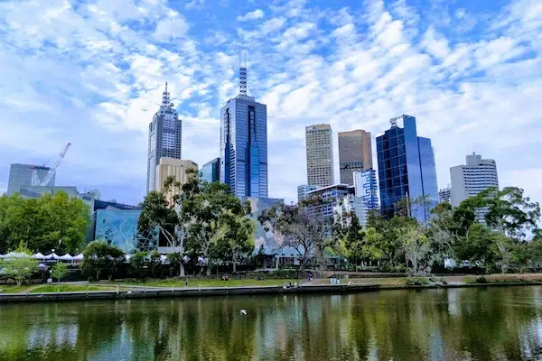 Melbourne city skyline with modern skyscrapers reflected in the Yarra River under a partly cloudy sky.