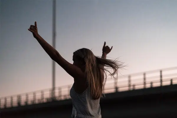 Silhouette of a woman with raised arms dancing at sunset on an urban bridge.