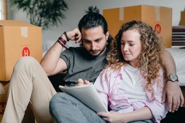 A couple sitting on the floor surrounded by cardboard boxes, reviewing a clipboard together during a move.