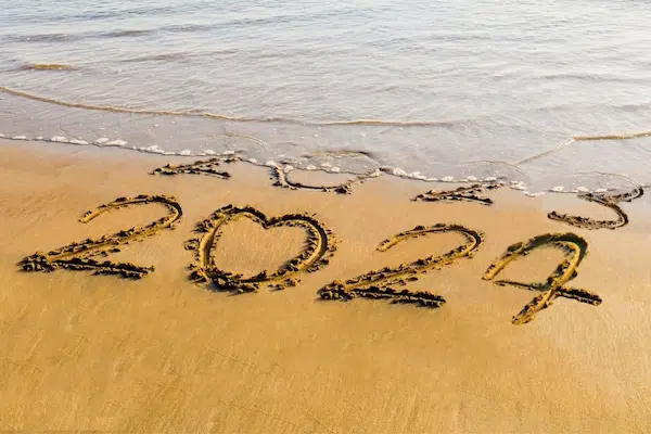 "2024" written in wet sand with a heart-shaped zero, as ocean waves begin washing away the years "2022" and "2023" above it.