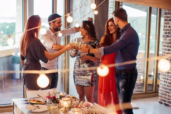 A group of five smiling friends, dressed up for a party, stand and toast with wine glasses under warm string lights.
